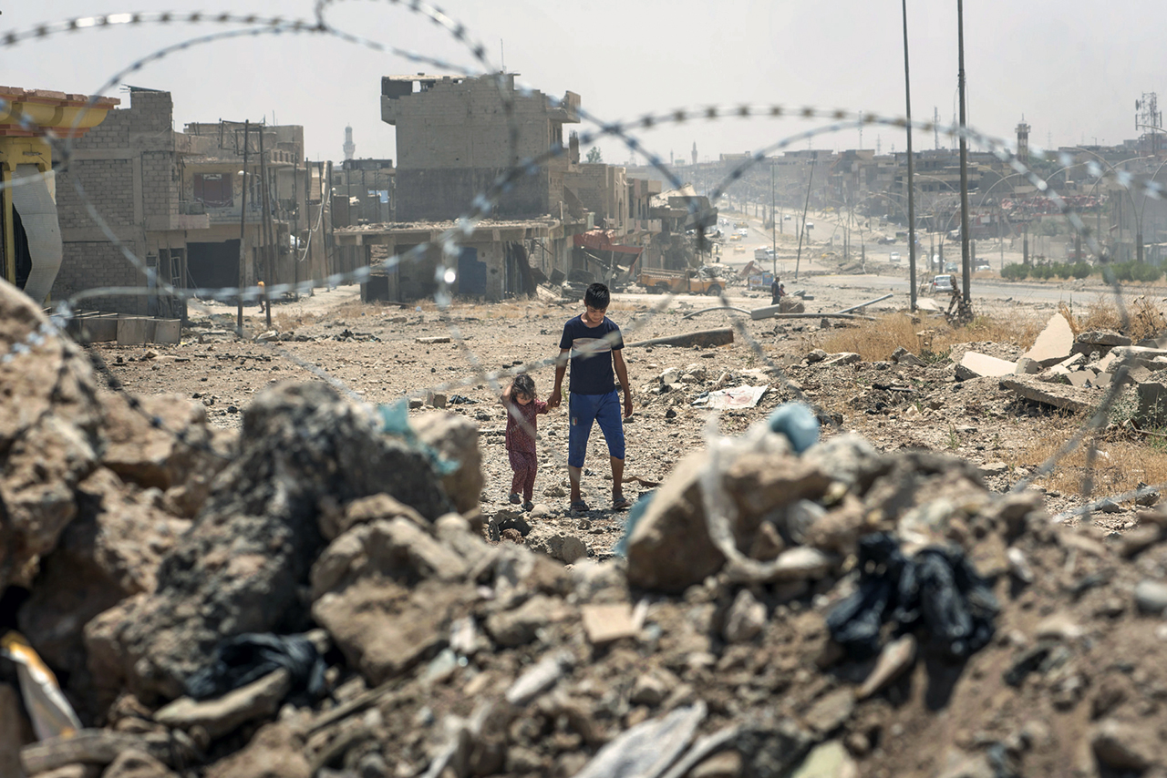 Two children walk amongst rubble as seen through a barbed wire fence