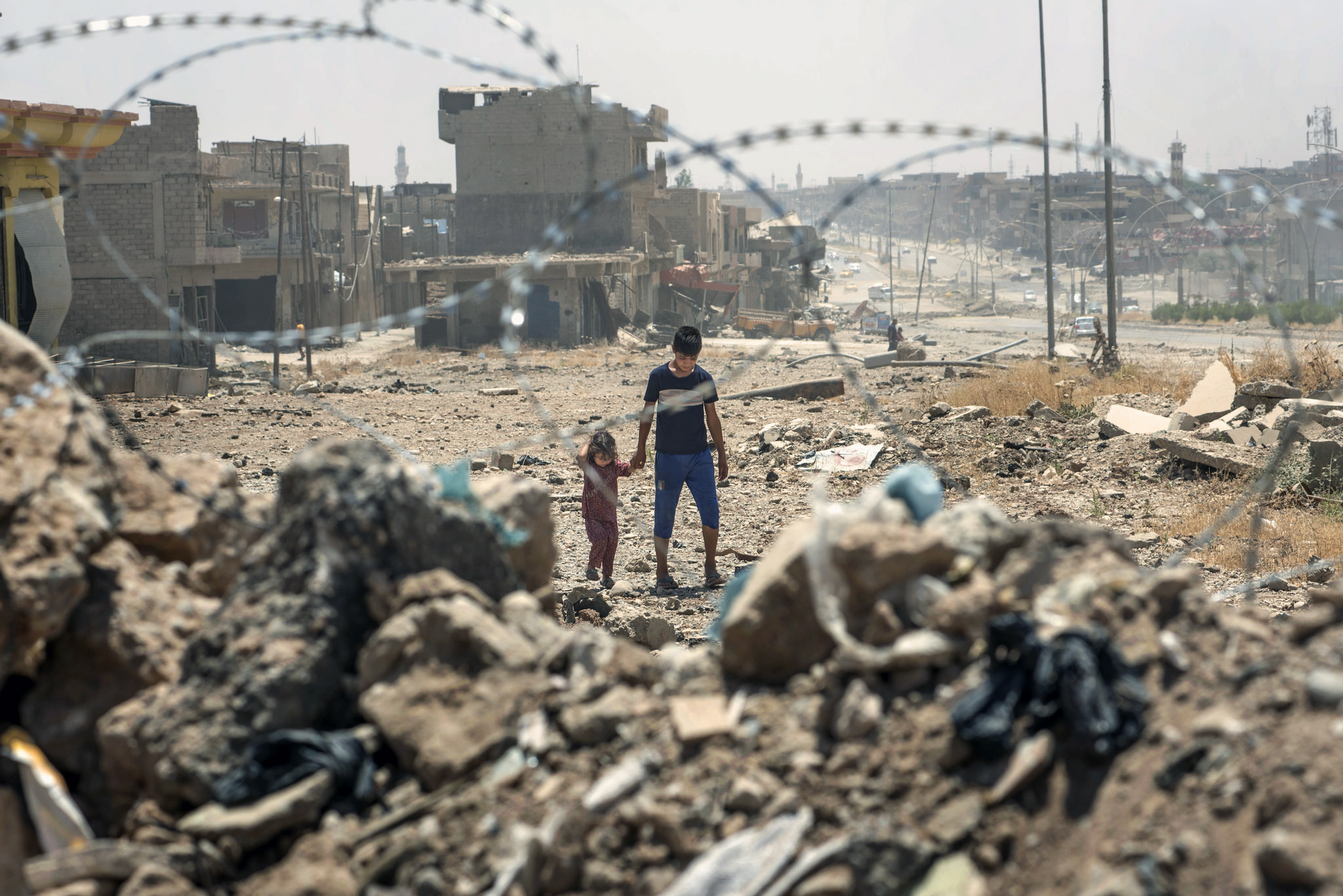 Two children walk amongst rubble as seen through a barbed wire fence