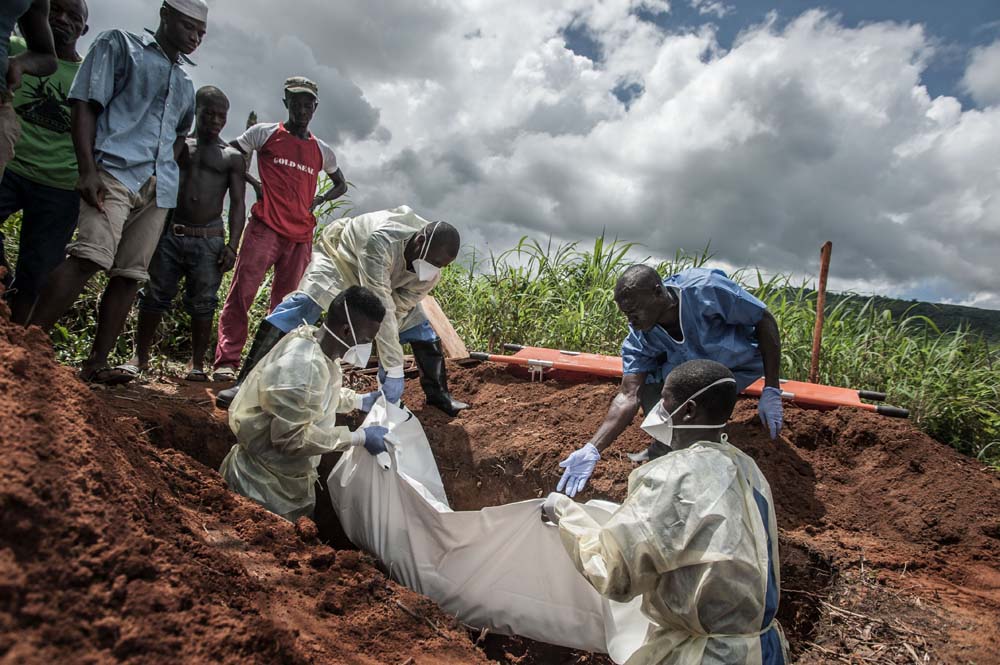 A group of burial workers in protective gear lower a covered body into a grave as onlookers stand by.