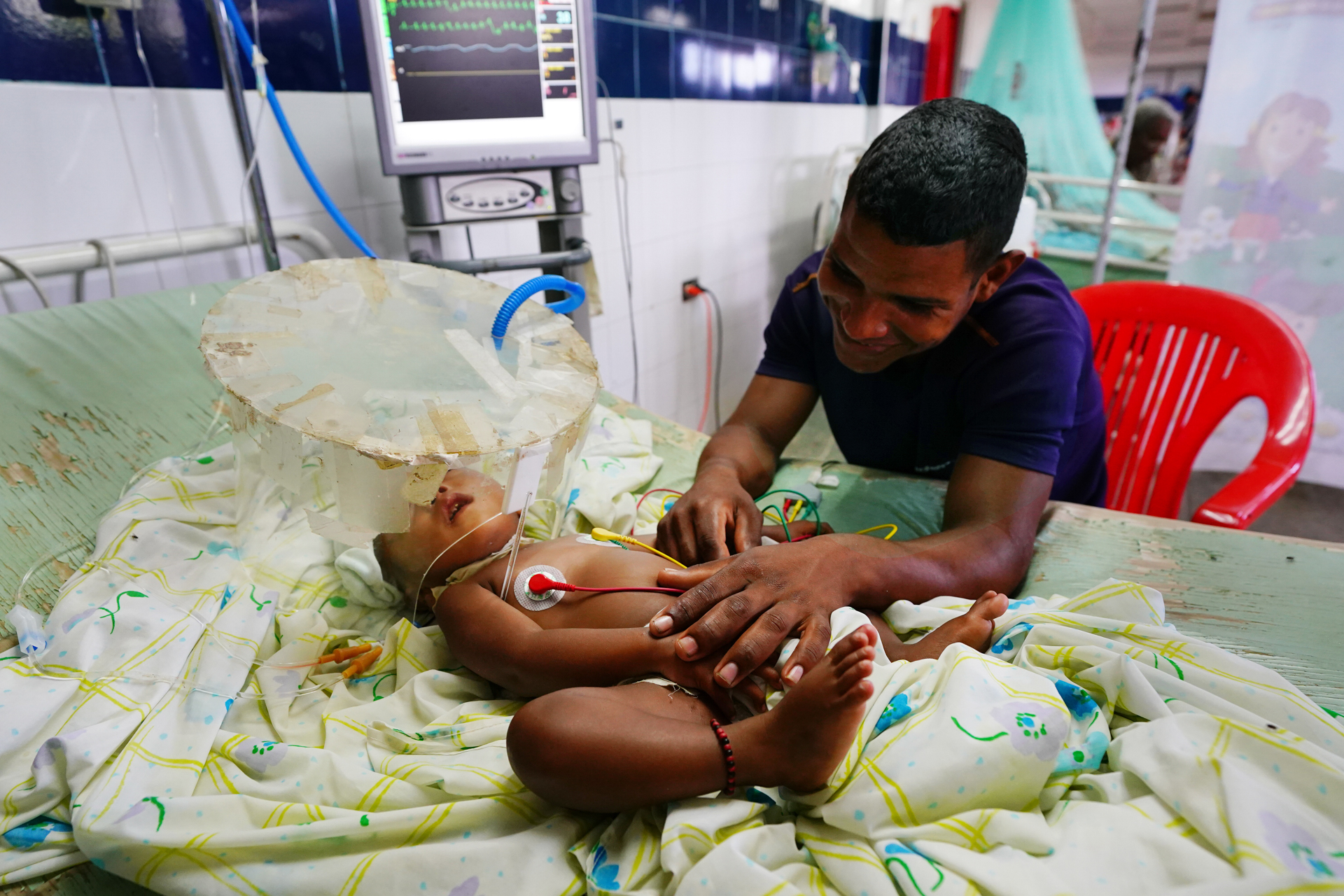 A man smiles over an infant in a hospital bed connected to equipment