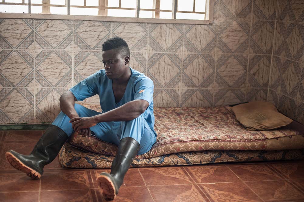 Tamba Momorie, an ebola burial worker in Sierra Leone, sits on a mattress on the floor in his scrubs.