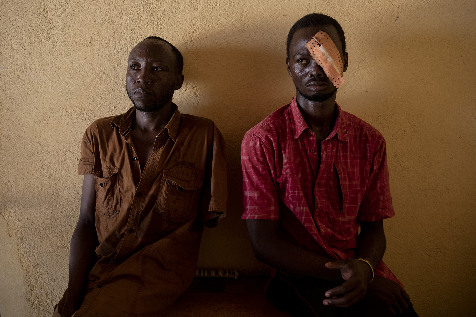 Two men sit on a wooden bench up against a cement wall. One of them wears a patch over his left eye.
