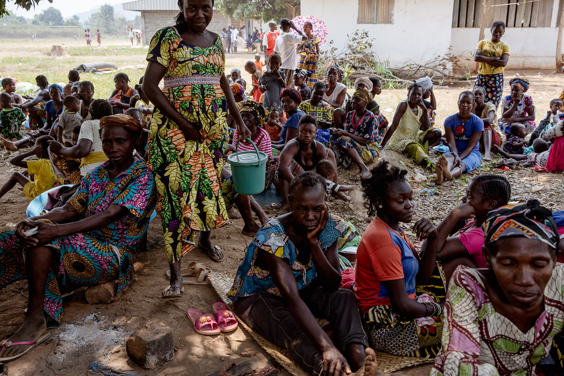 A group of villages of all ages fill the frame, sitting on the ground in a displacement camp.