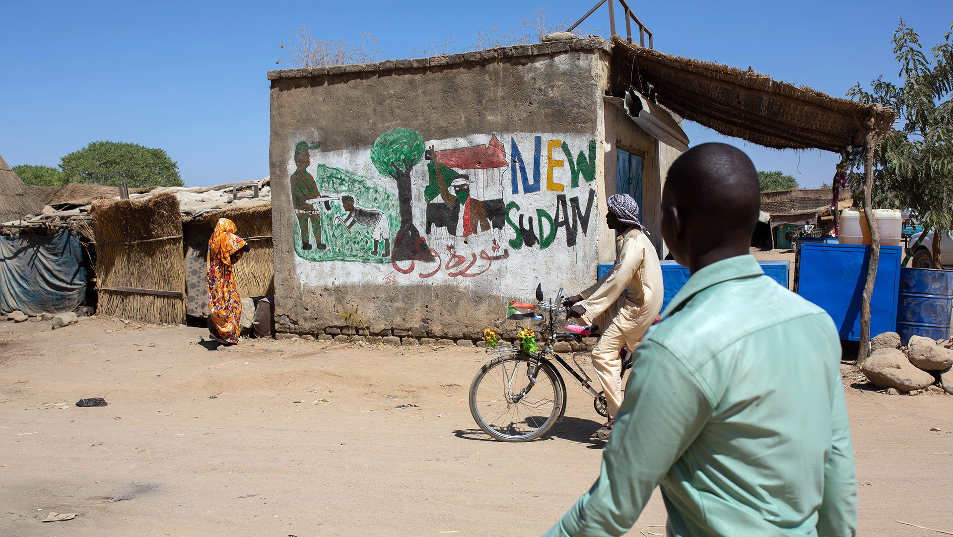 A man walks down a dusty street, past a painted mural.