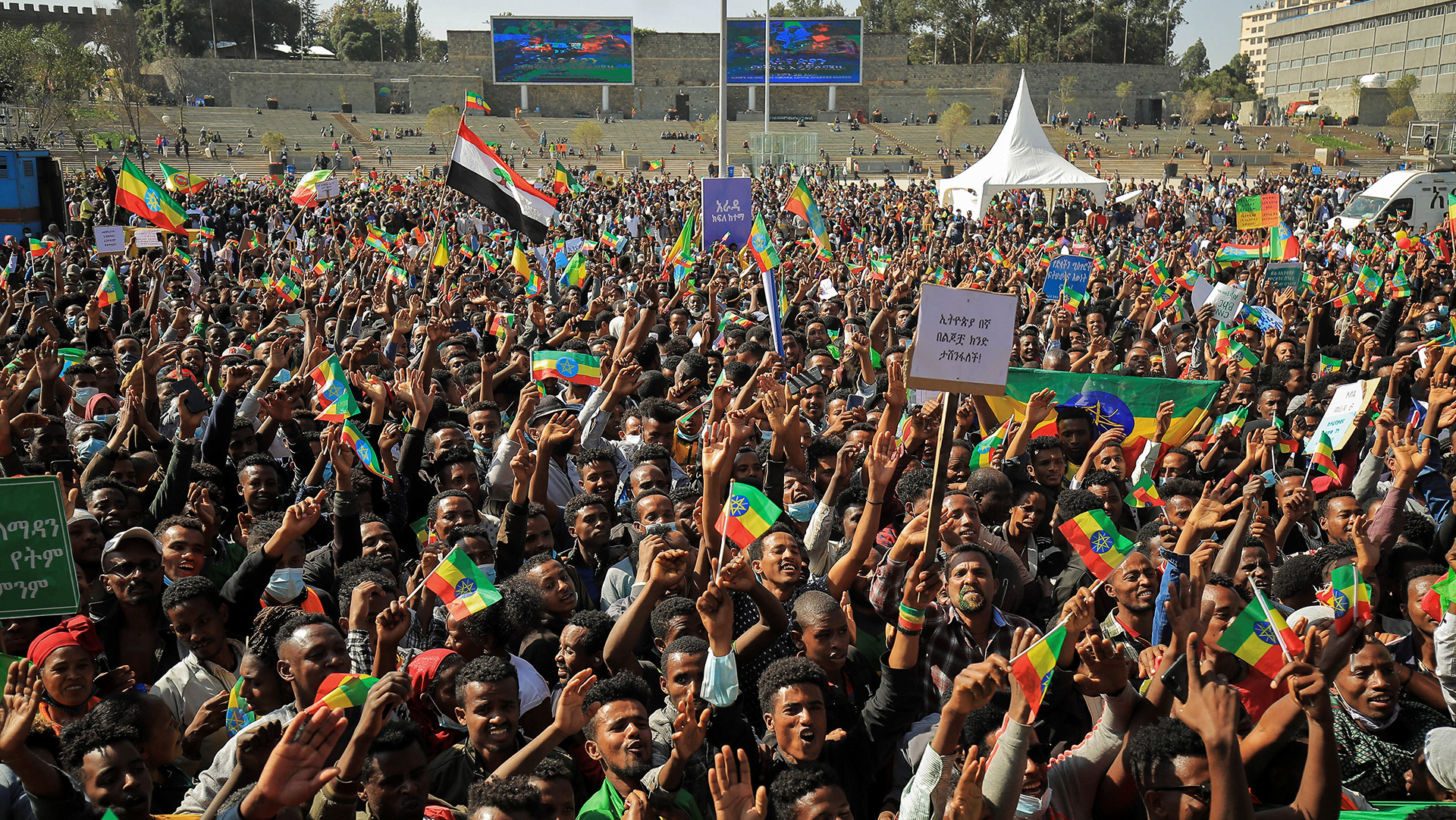 A large crowd of people gathered in a city square are holding flags and shouting