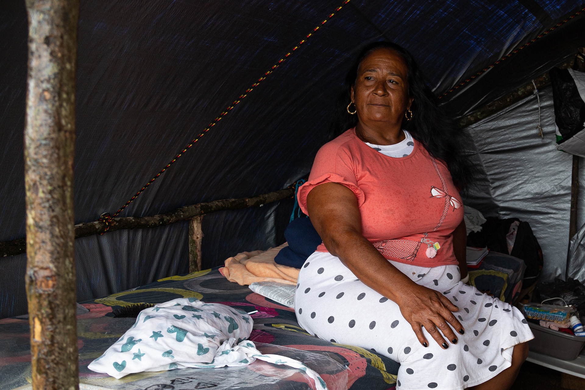 A cuban migrants in a brightly coloured shirt sits on a bed in her makeshift shelter looking past the camera.