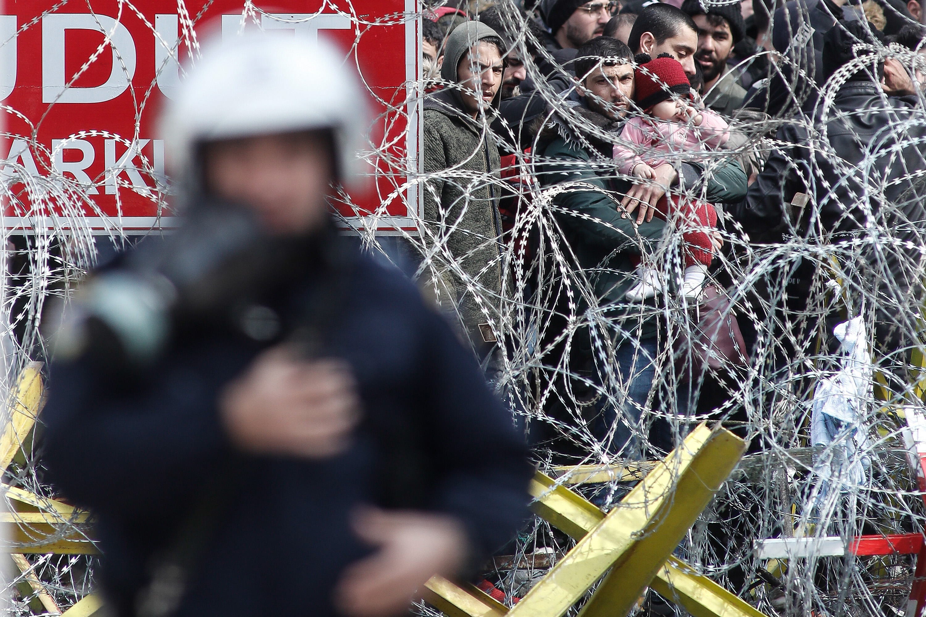 A photo of Greek riot police patrol along a barbed-wire fence. Asylum seekers gather on the other side of the fence.