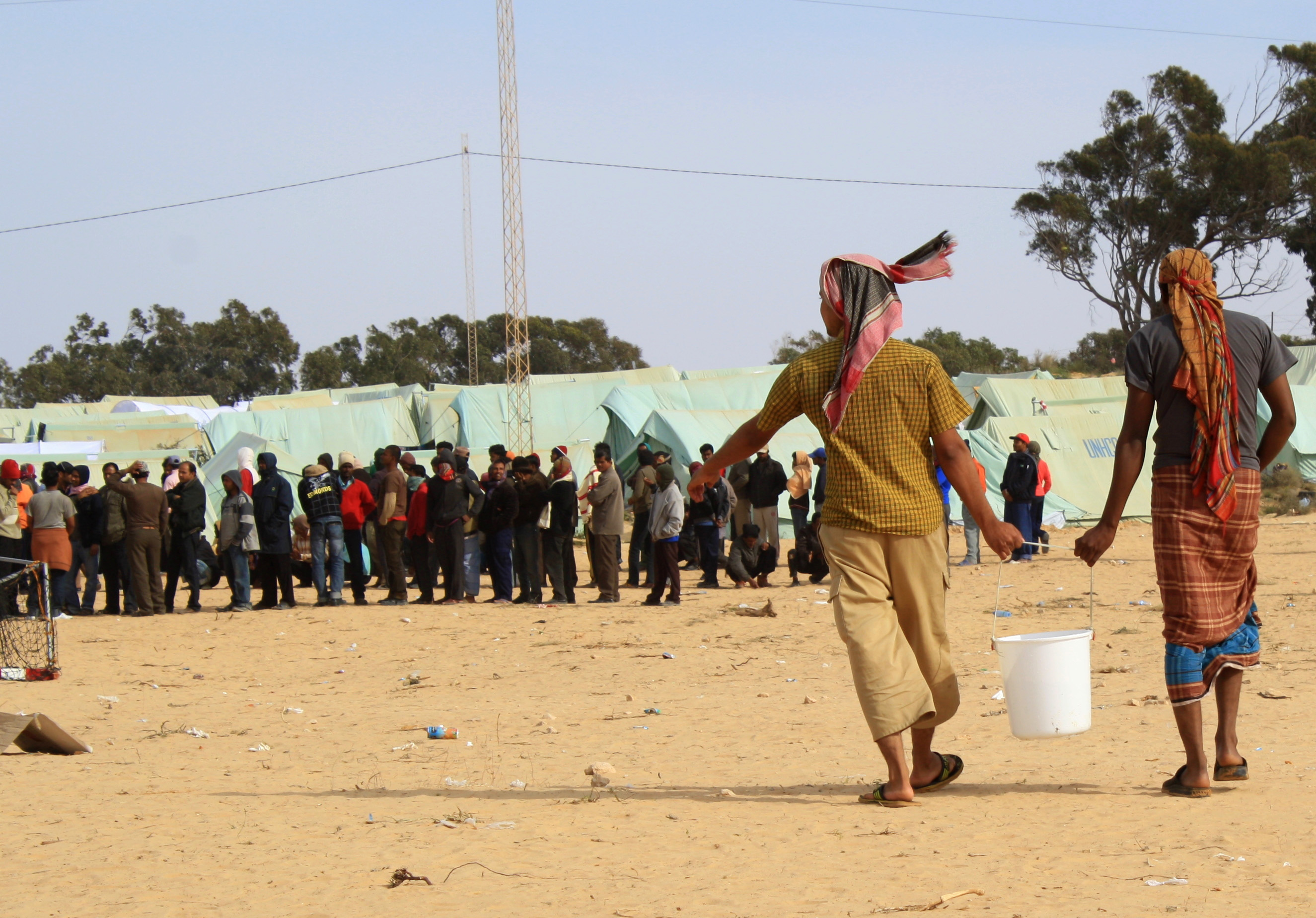 Men carry a bucket of water at Choucha camp whilst others queue for assistance in the background