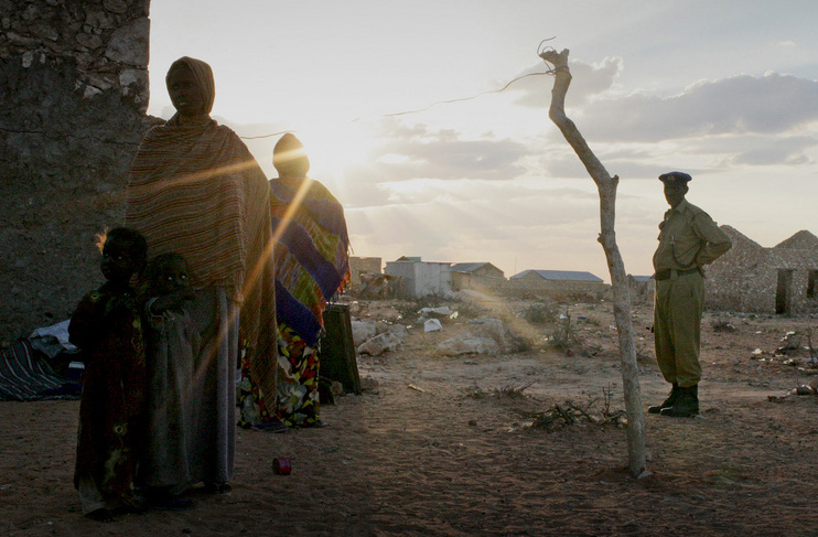 A woman and her young children stand in the evening light at an IDP settlement in South Galkayo, Somalia with armed police standing by