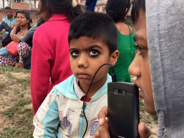 Children living in a temporary camp listen to the radio on a smart phone after being displaced by a 7.8 magnitude earthquake that struck Nepal 25 April 2015