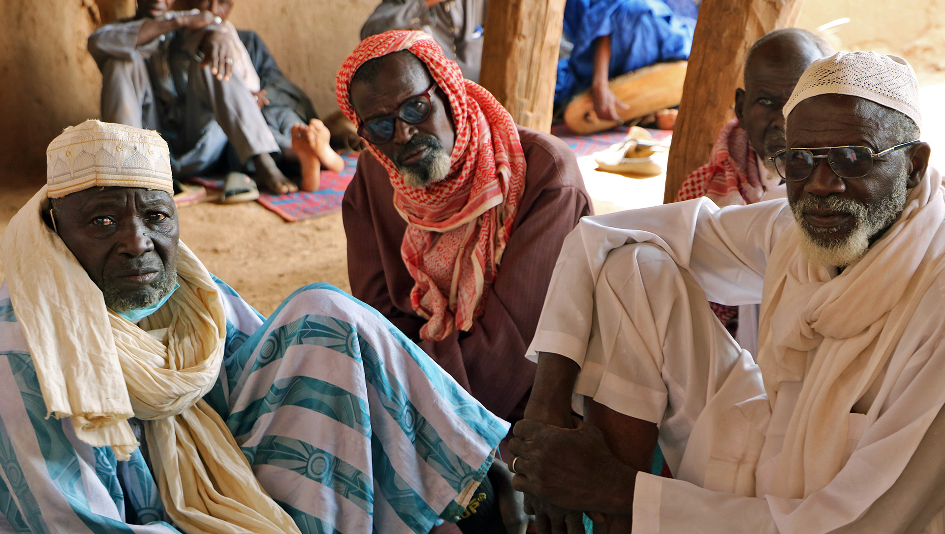 Three men sit on the ground under a shelter, looking up at the camera.