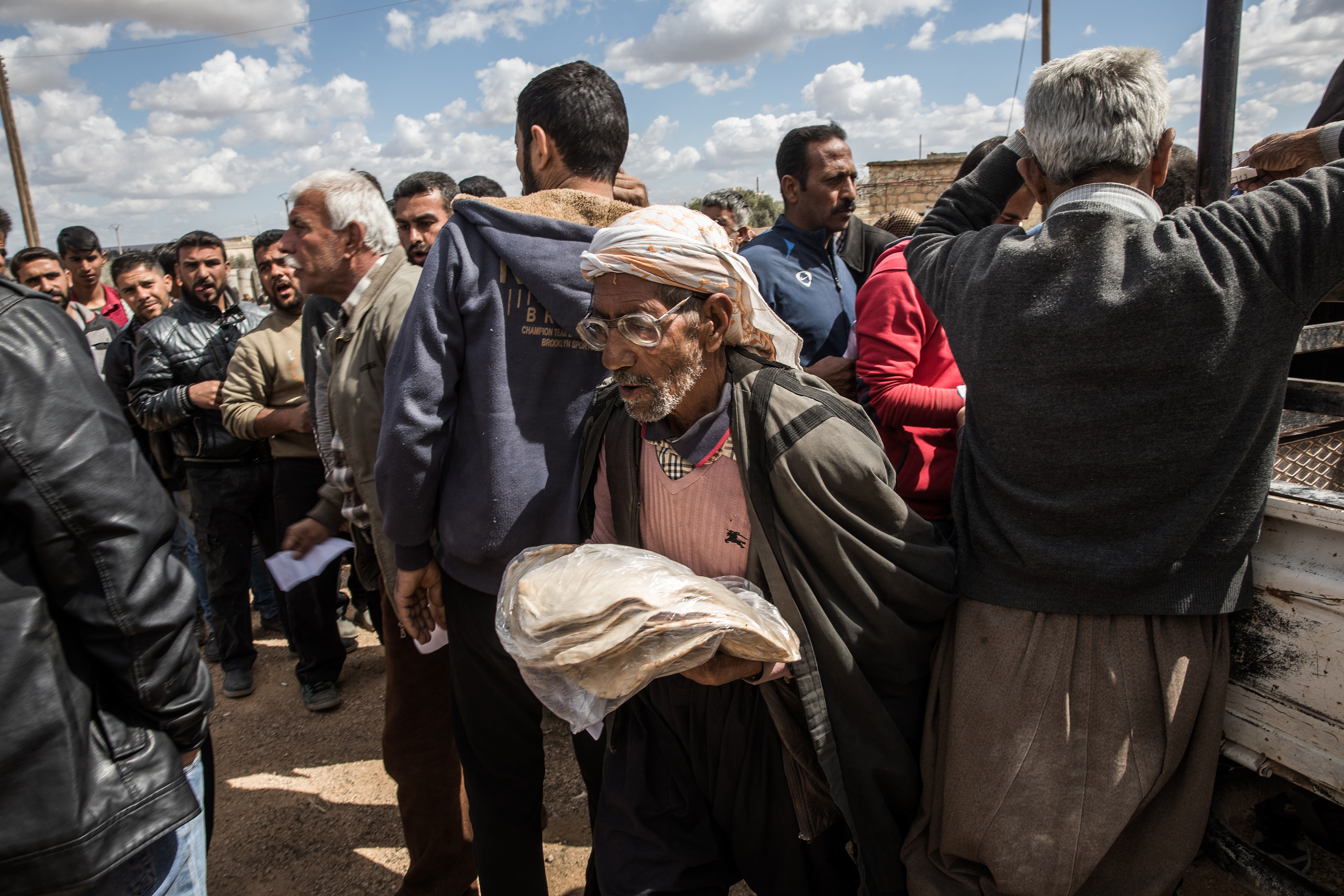 A crowd gathers around a truck. An older man, the focal point of the photo, walks away with bread in hand.