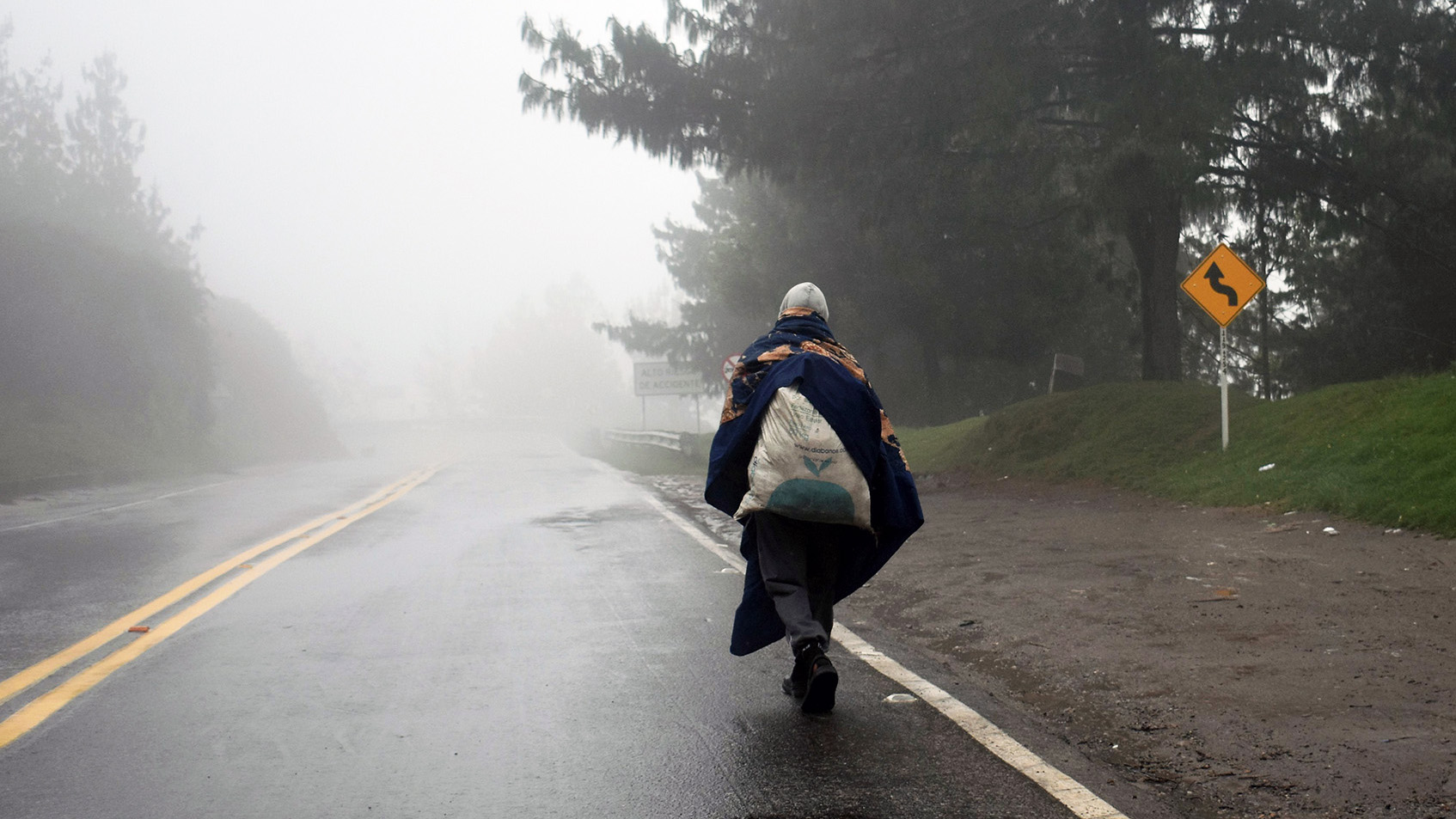 A figure walks along a foggy highway, a bad slung over their shoulder.