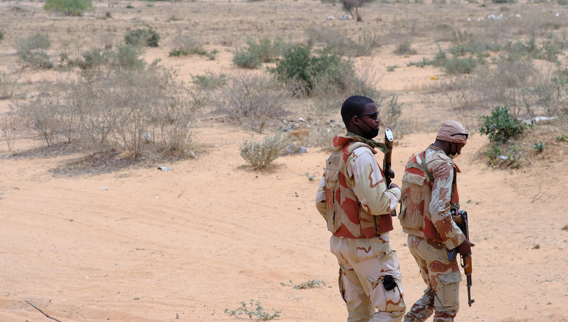 Two soldiers walk off of a dusty road into brush land.