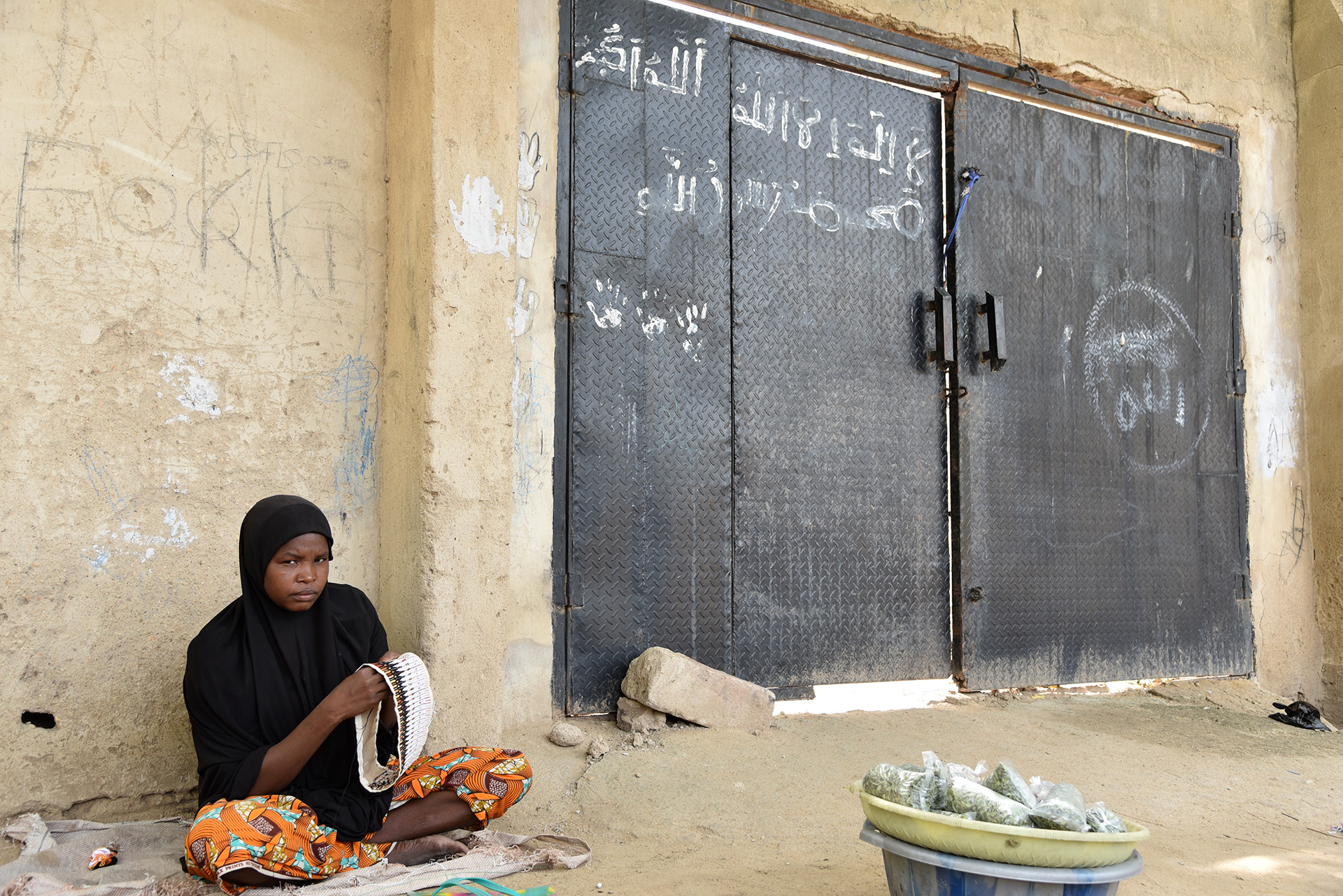 A woman sews caps for sale in Bama, next to a gate daubed with Boko Haram graffiti: “There is no God but Allah”. Boko Haram was pushed out of the town in 2015.