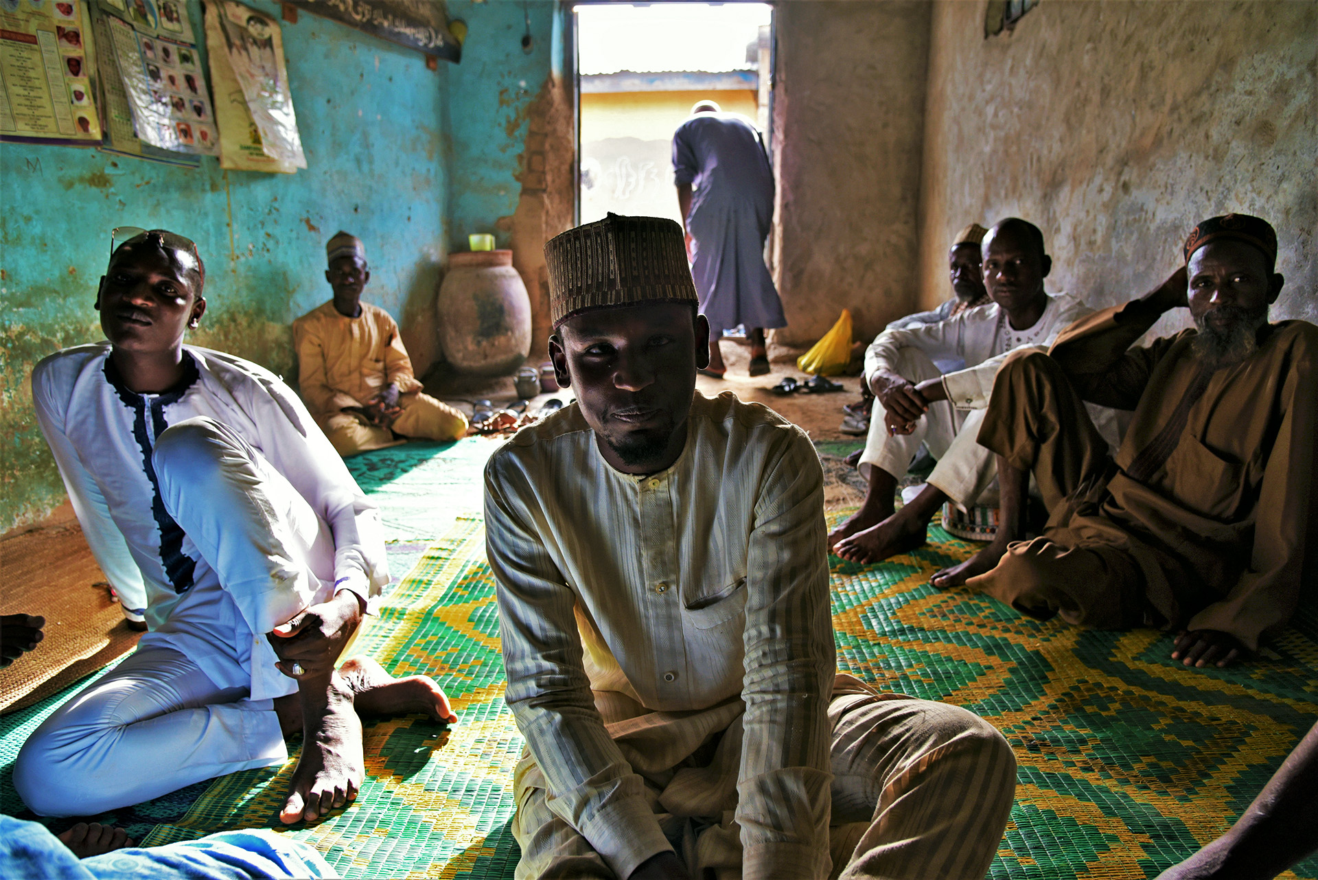 Members of the Miyetti Allah Cattle Breeders Association sit in a sparsely decorated room, sunlight streaming in through the door behind them.