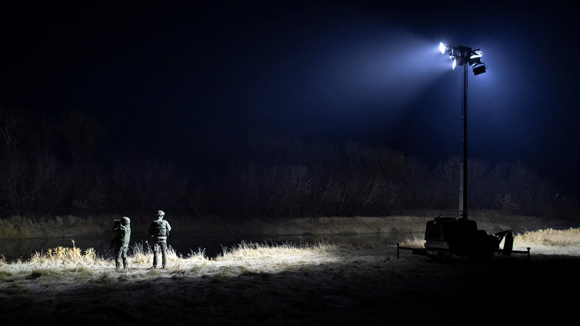 Two guards stand illuminated by a spotlight at night in a forested area.