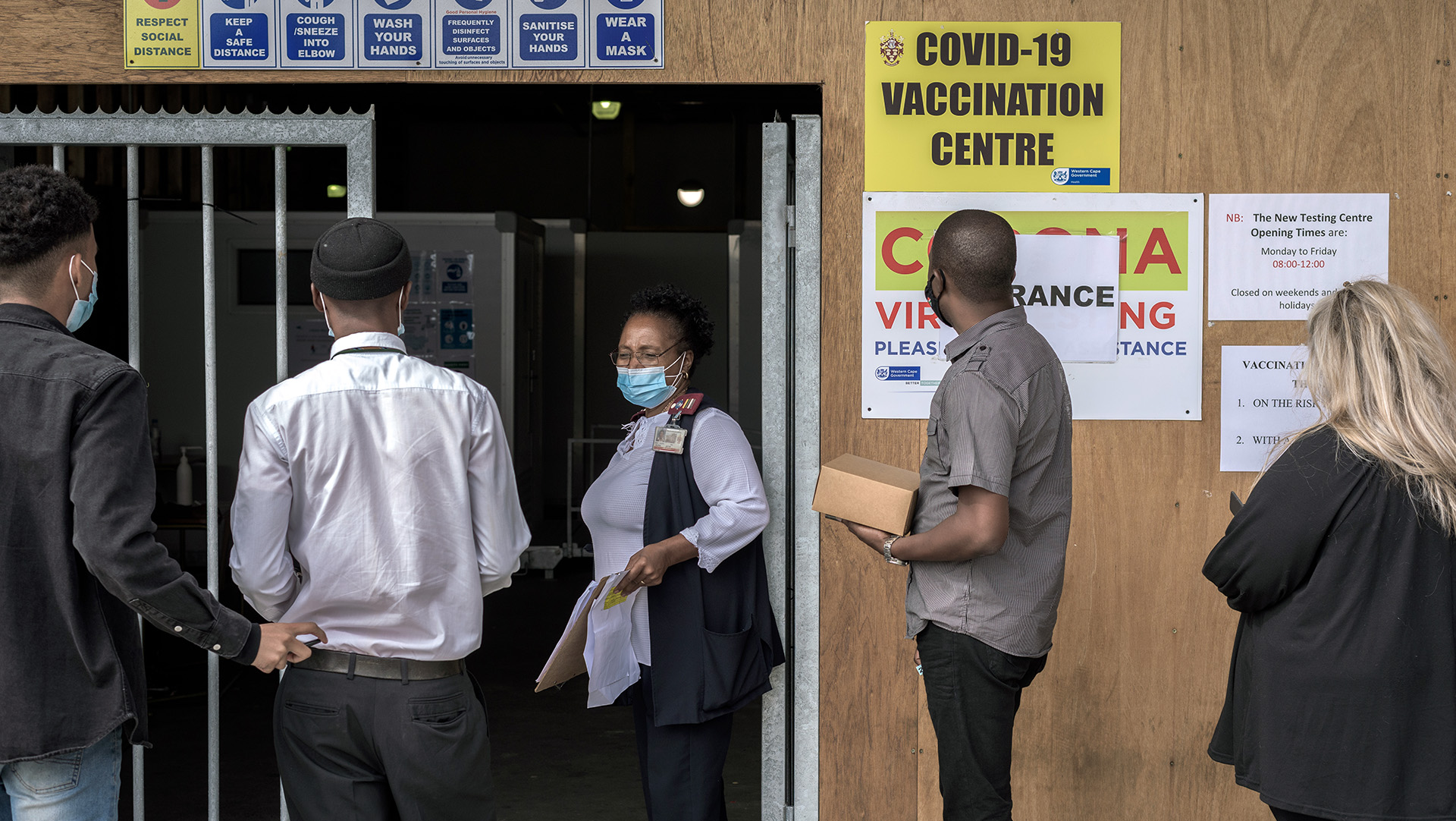 A number of people line up in front of a health centre, their backs facing the camera