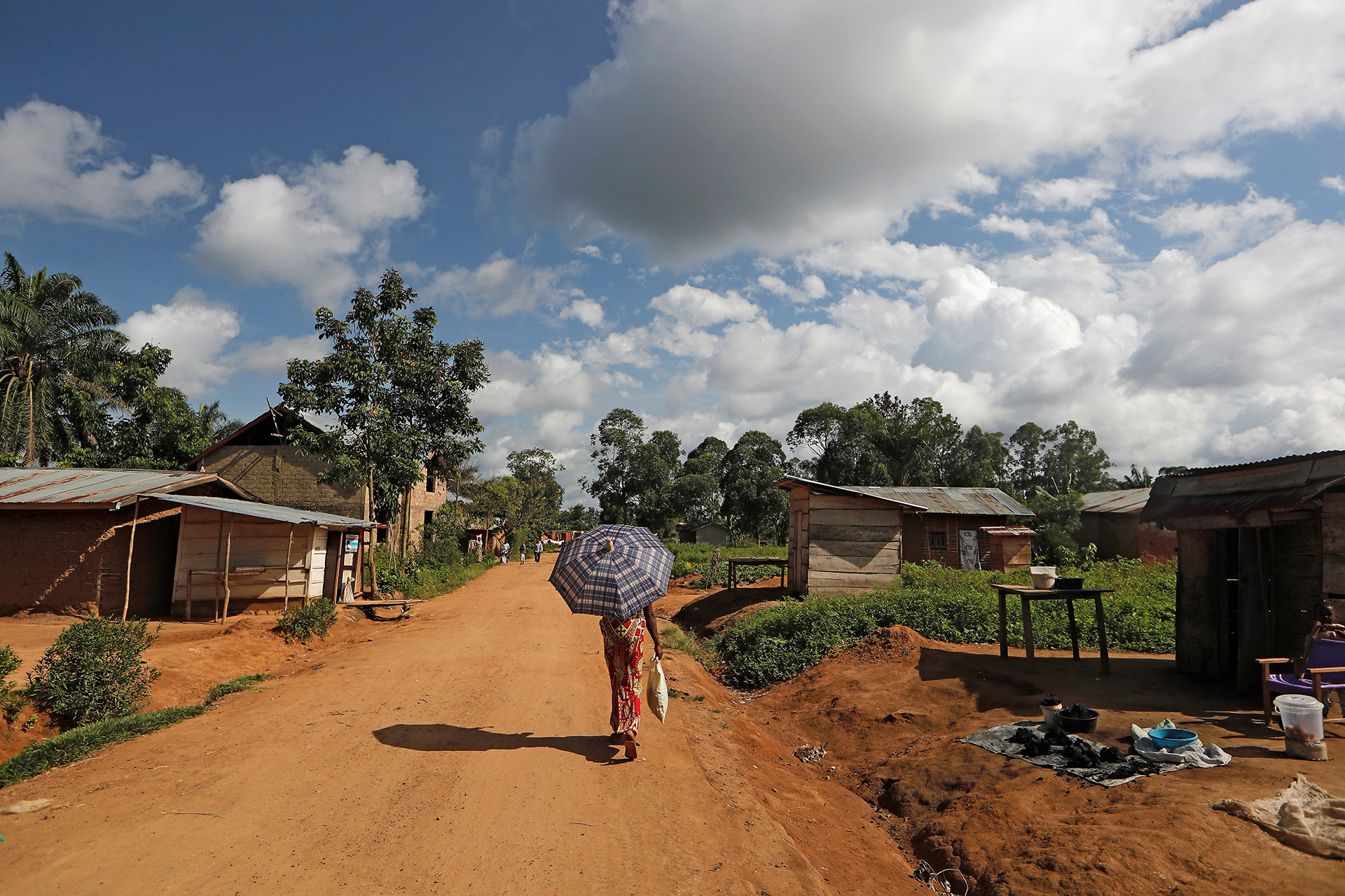 A woman walks down a dirt road away from the camera, holding an umbrella.