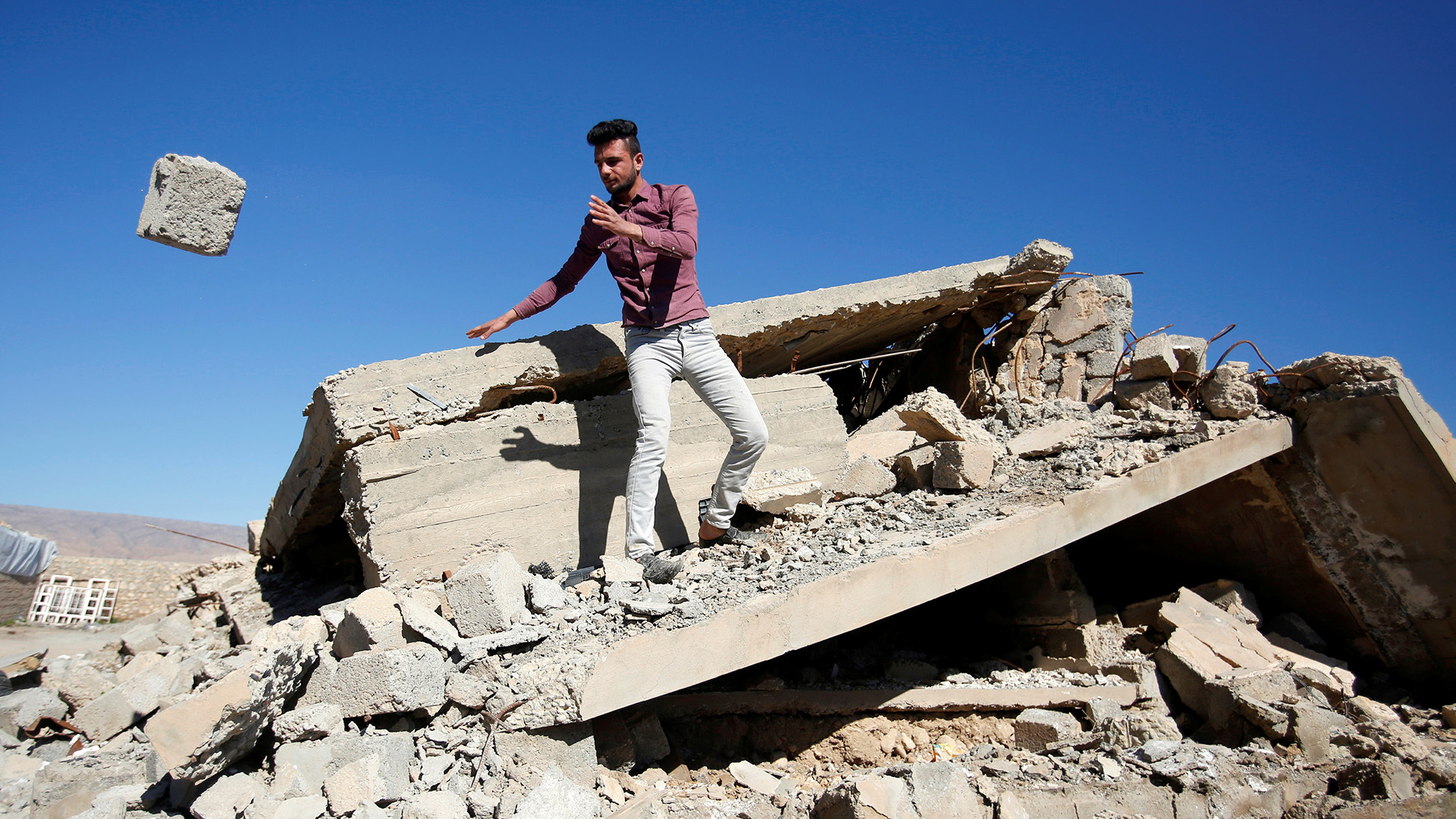 A man stands on a pile of rubble, a large stone he has thrown is in mid-air
