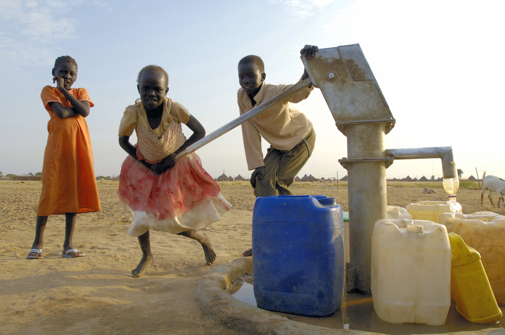 Children are looking at the camera and jumping on the handle to a water pump