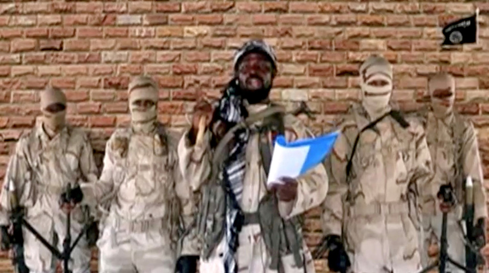 The leader of Boko Haram stands with a gun wearing fatigues in front of a green screen brick wall.