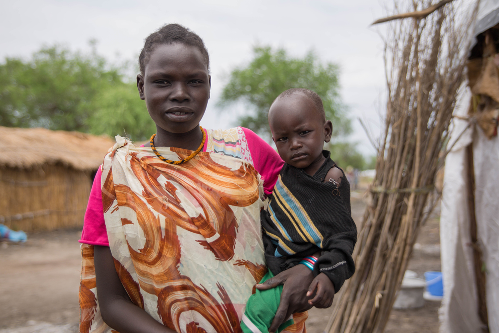 A young woman with a child on her hip in multi-patterned fabric looks at the camera