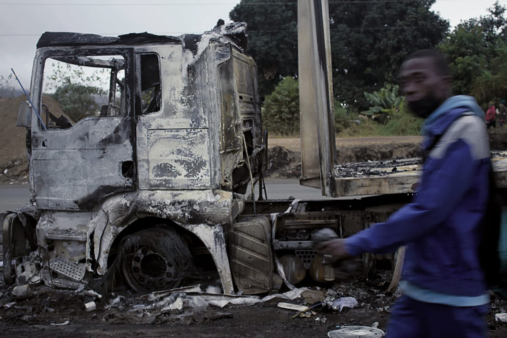 A man walks in front of a burnt out truck.