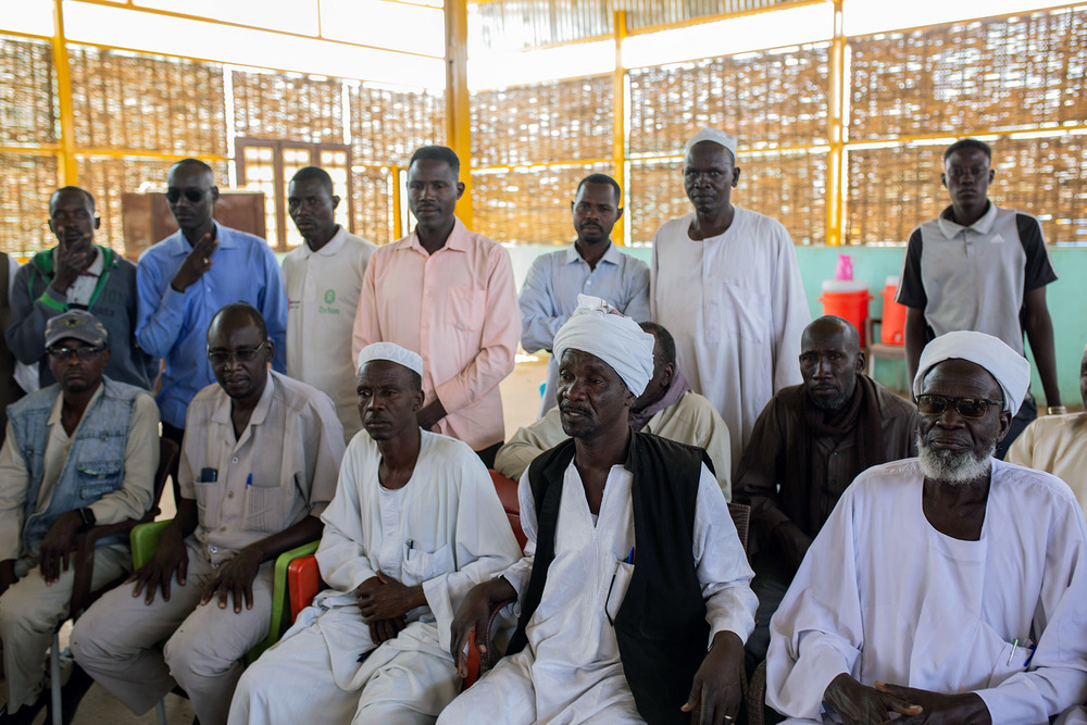 Men stand and sit in chairs in a covered shelter
