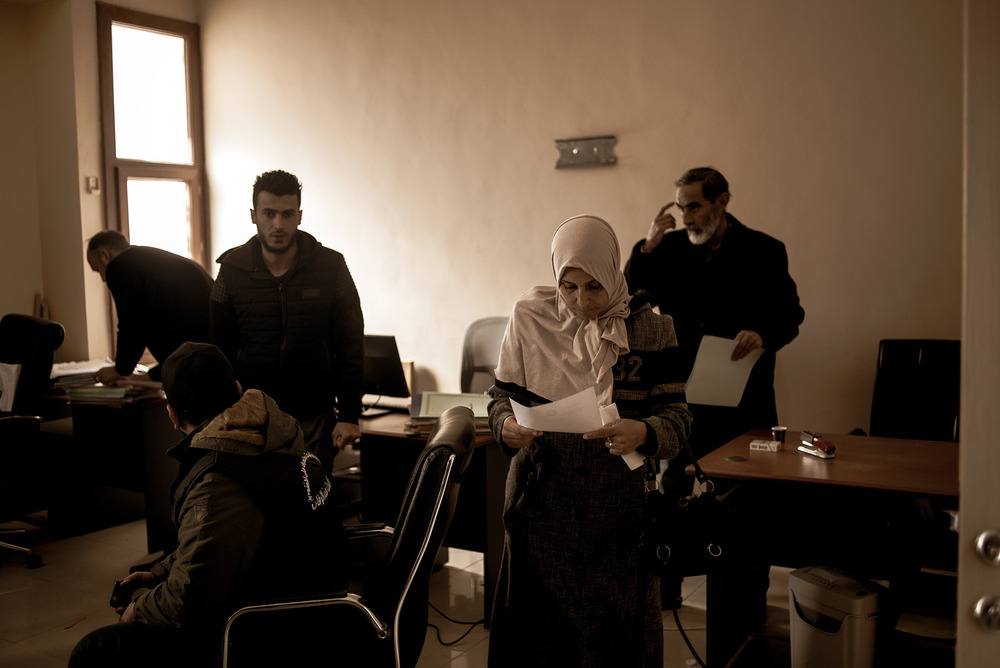 A woman in a headscarf walks out of an office room holding a document in her hand – several men are seen in the background.