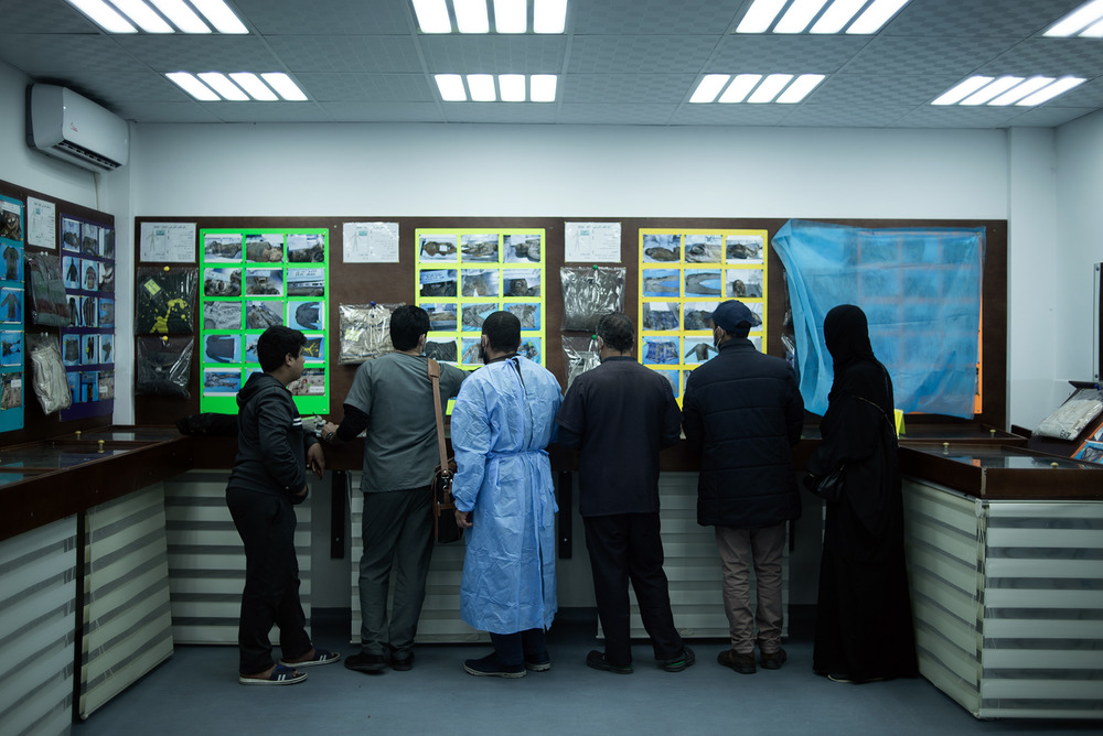 The image shows a line of people looking at pinned images on a hospital wall, their backs to the camera.