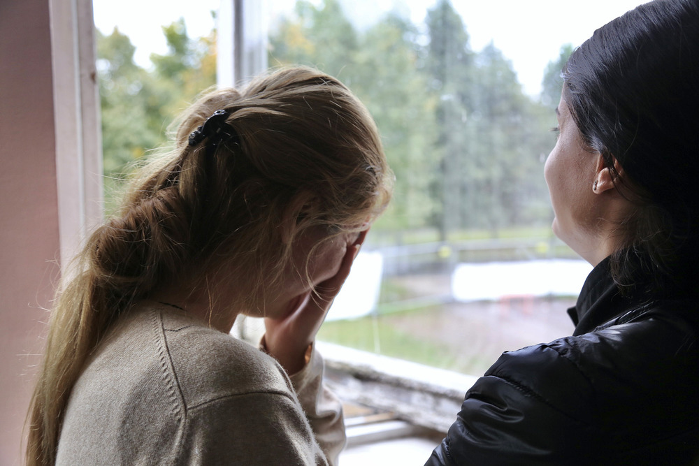 A woman stands facing a window, holding her head in her hands.