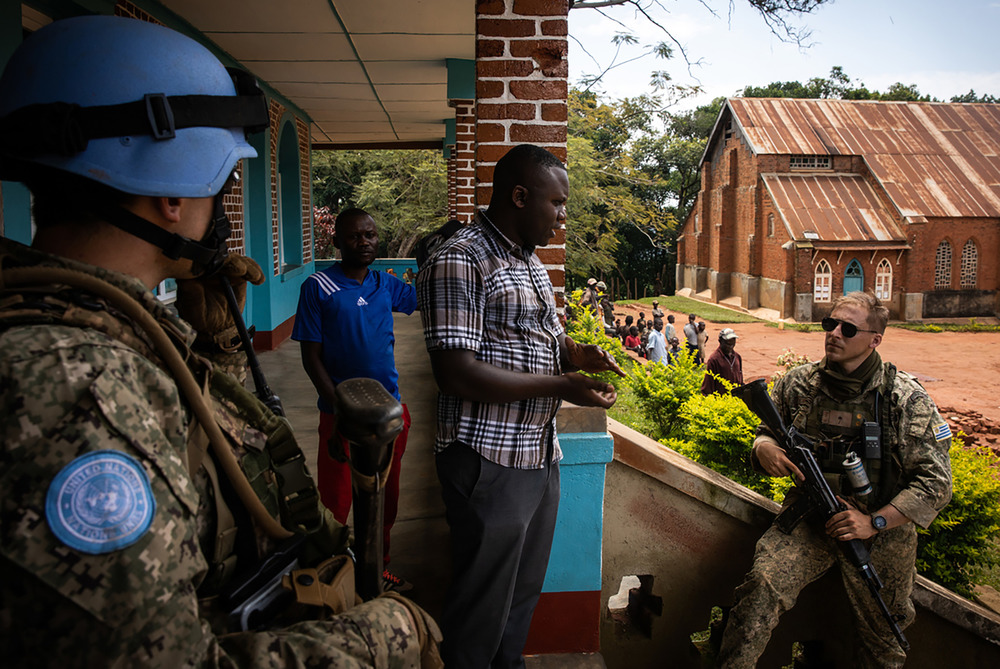 A priest stands on the steps of a building speaking with an armed peacekeeper.