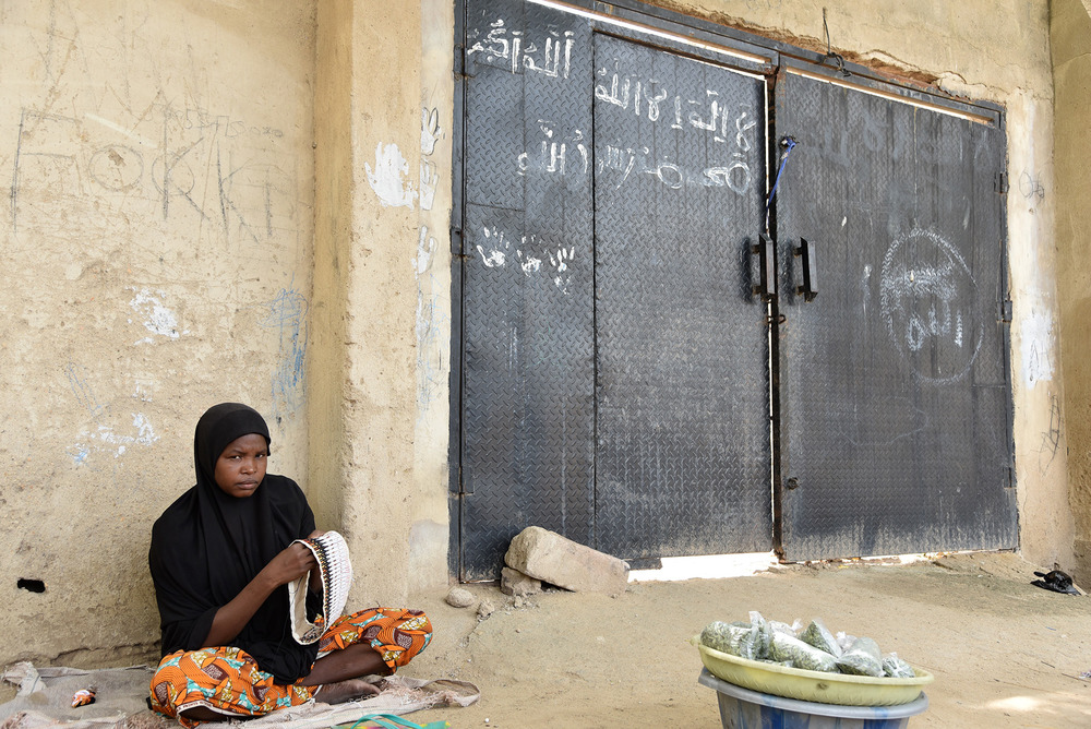 A woman sews caps for sale in Bama, next to a gate daubed with Boko Haram graffiti: “There is no God but Allah”. Boko Haram was pushed out of the town in 2015.