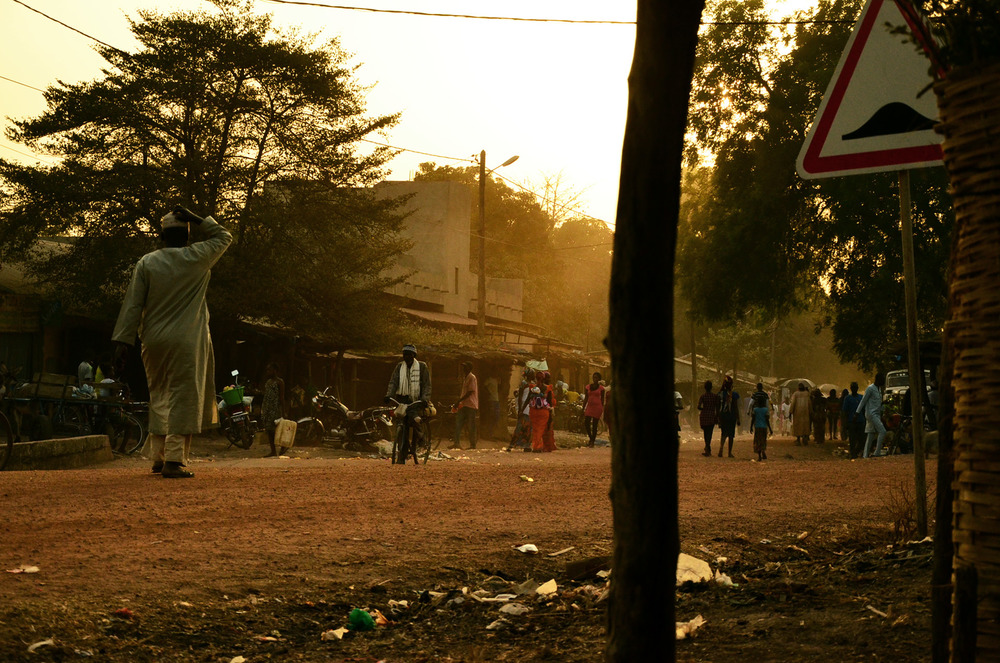 Several people walk down a dusty street in a village as the sun sets.