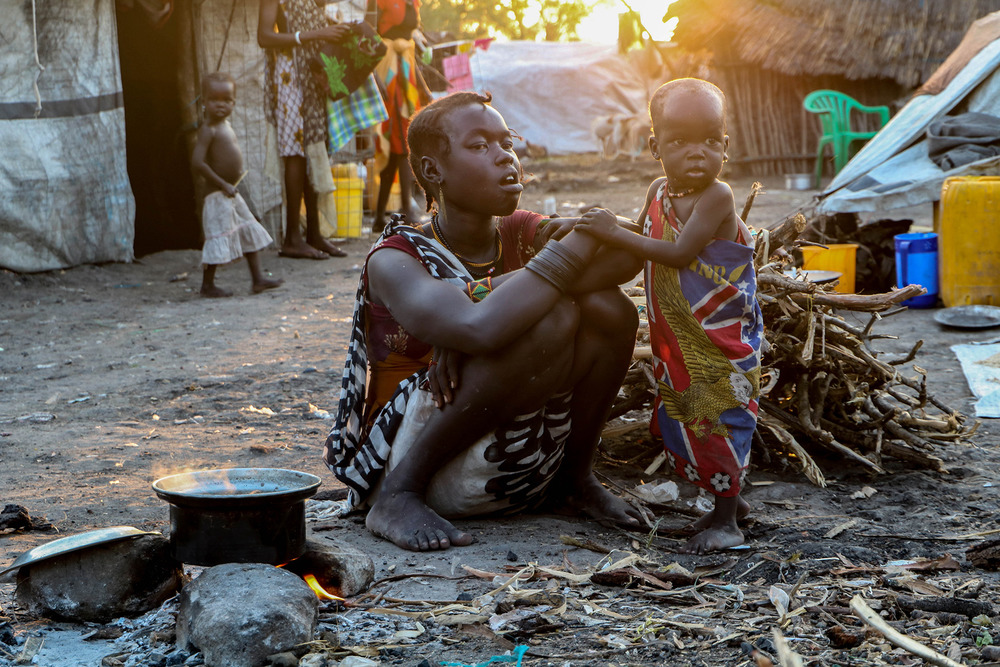 A woman crouches near her child beside a small pile of firewood in a makeshift displacement camp.