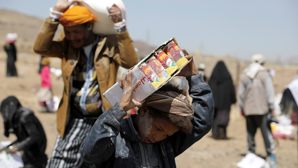 A youth and a man carry food aid on their shoulders in the sun.