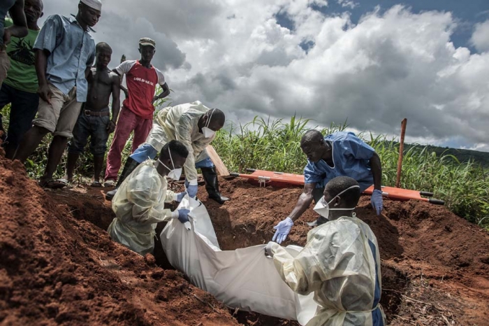 A group of burial workers in protective gear lower a covered body into a grave as onlookers stand by.