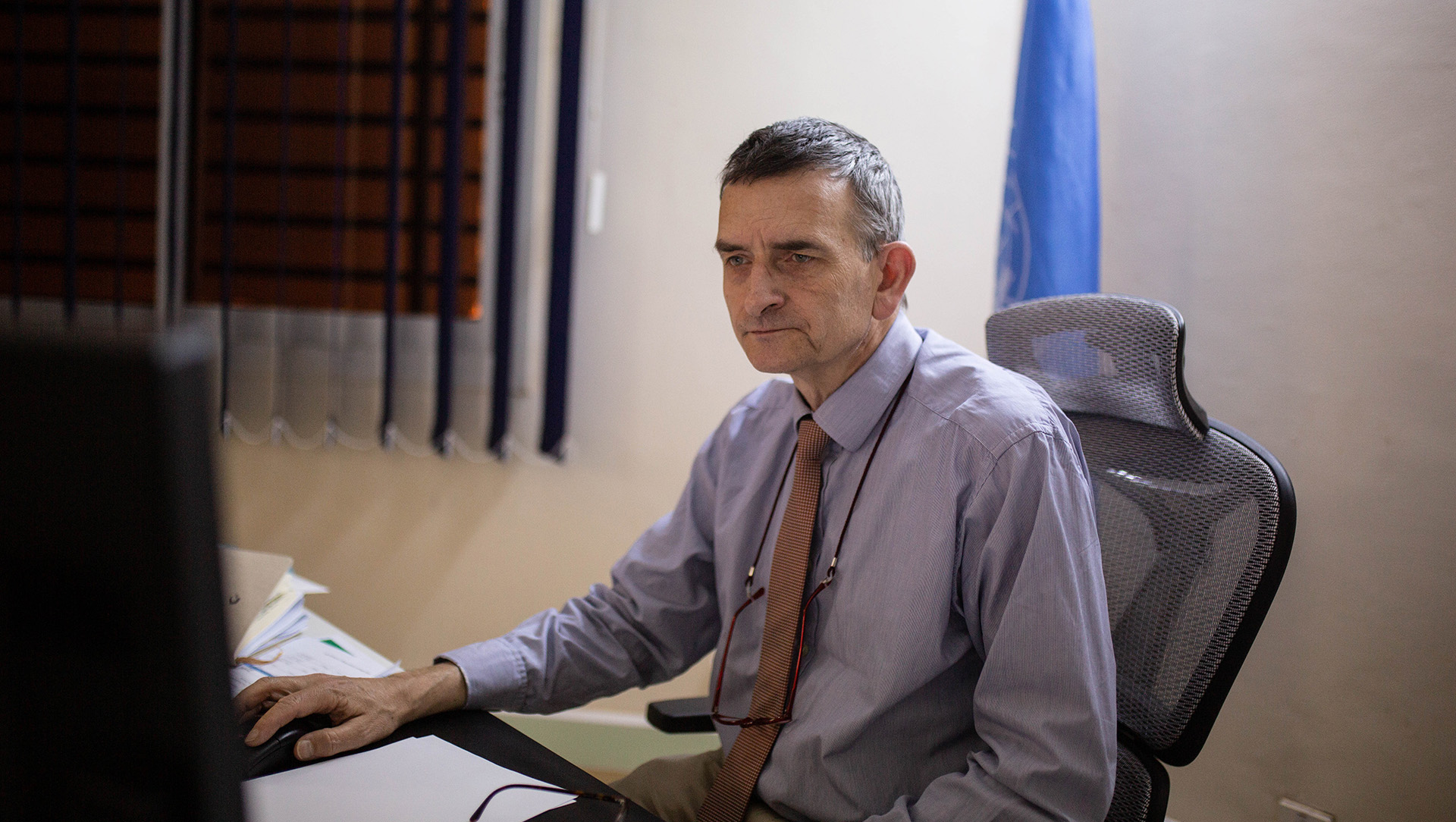 A man sits at a computer desk with a UN flag behind him.