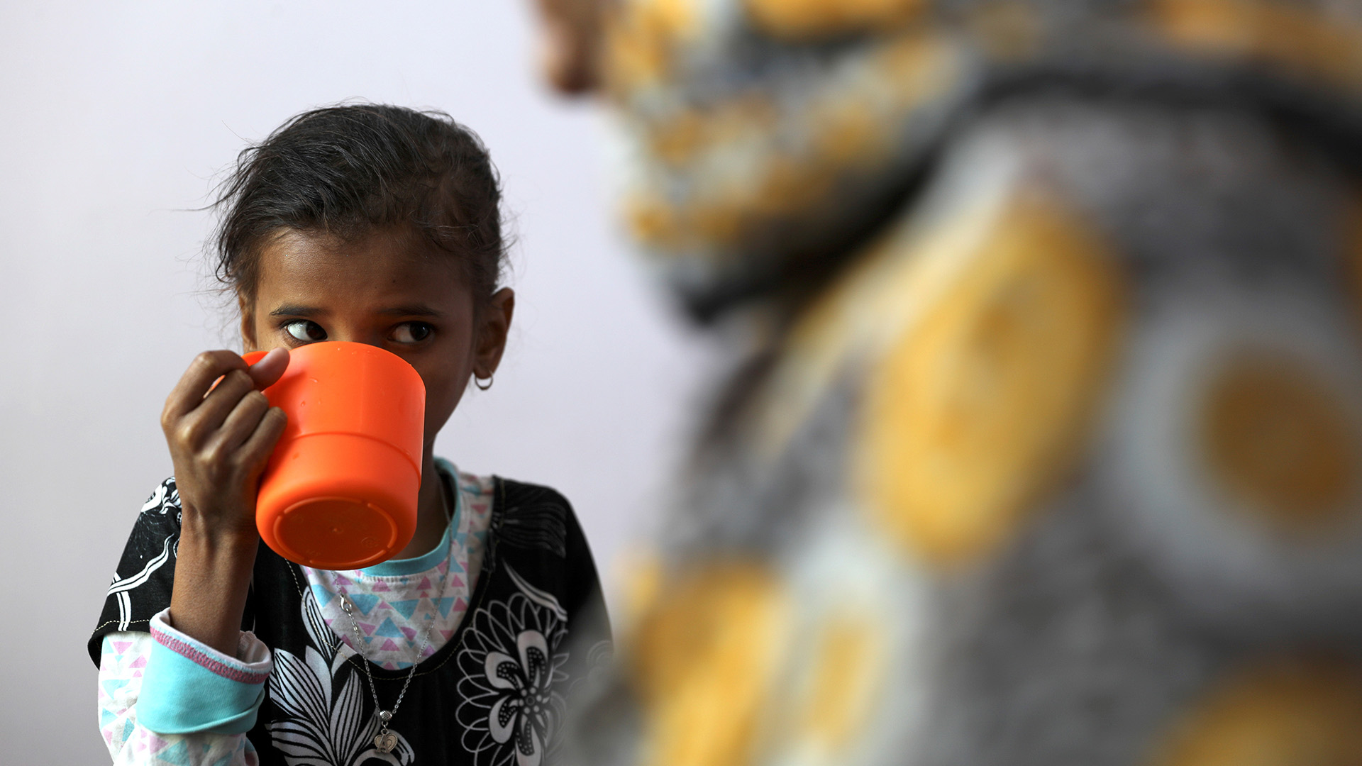A young girl drinks out of an orange plastic cup on the left hand side of the image, while of the right hand side the blurred outline of a figure fills the foreground