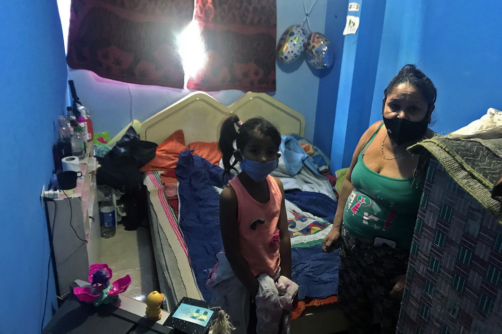 Adolia Silva stands with her granddaughter in front of a bed in a small blue painted bedroom, looking at the camera.