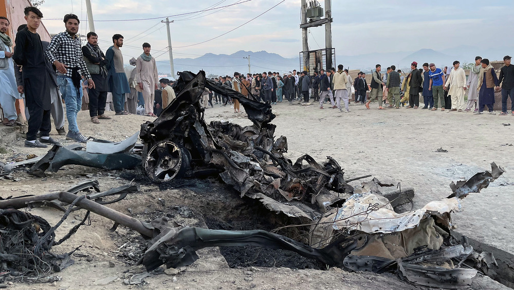 A crowd of people stand around the wreckage of a car in the sandy plaza of a city