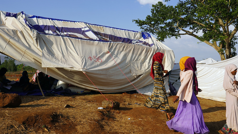 Several women walk to the right of the frame, behind them a makeshift shelter in a desert-like landscape.