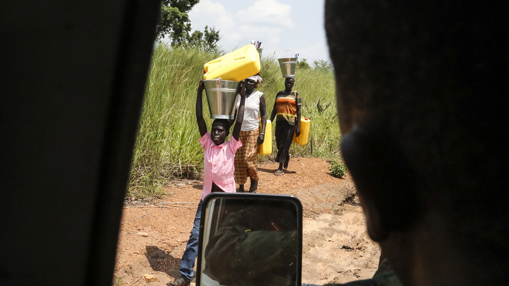 Several refugees walk along the side of the road, carrying large jugs of water on their heads.