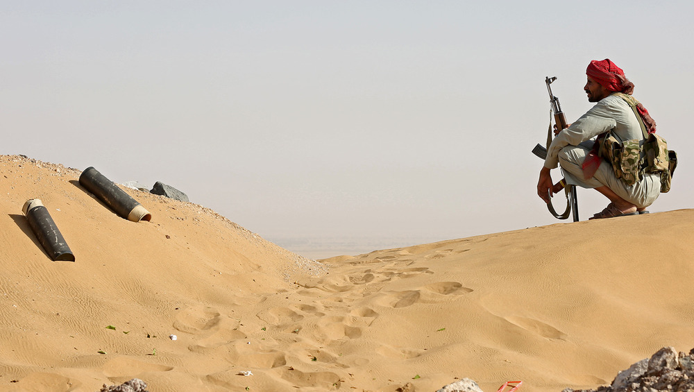 A man crouches on a sand dune, holding a rifle