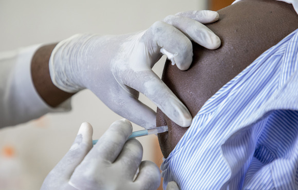 The image shows the upper arm of a patient being injected with a vaccine.
