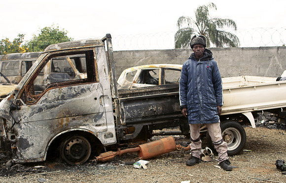 A man stands beside a burned out car.