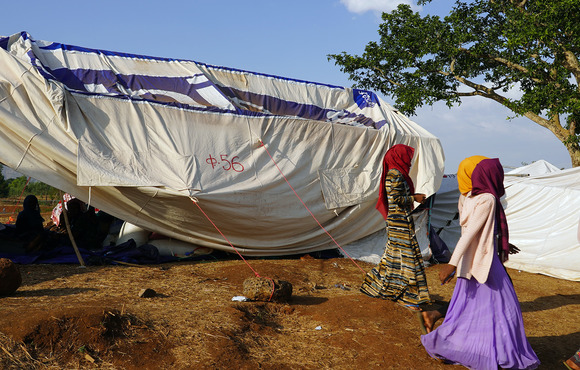 Several women walk to the right of the frame, behind them a makeshift shelter in a desert-like landscape.