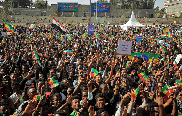 A large crowd of people gathered in a city square are holding flags and shouting
