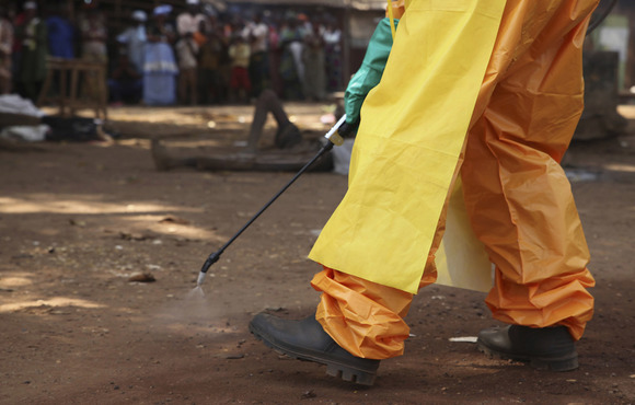 The image shows the bottom half of a person's body - dressed in a hazmat suit, apron, gloves, and boots - holding a disinfectant wand spraying the ground. 